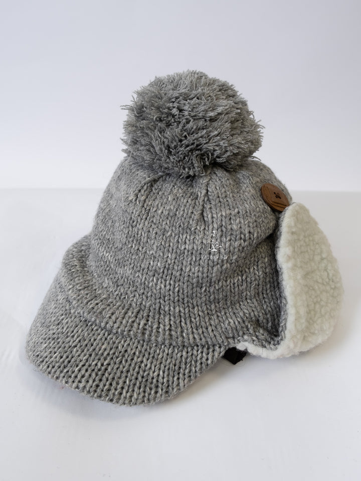 Grey knit trapper cap with large pom-pom and soft sherpa earmuffs, shown from a front-side angle.